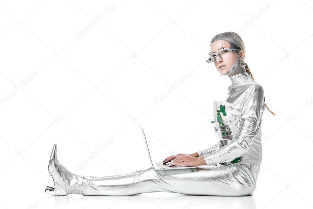 side view of silver robot sitting with laptop and looking at camera isolated on white, future technology concept