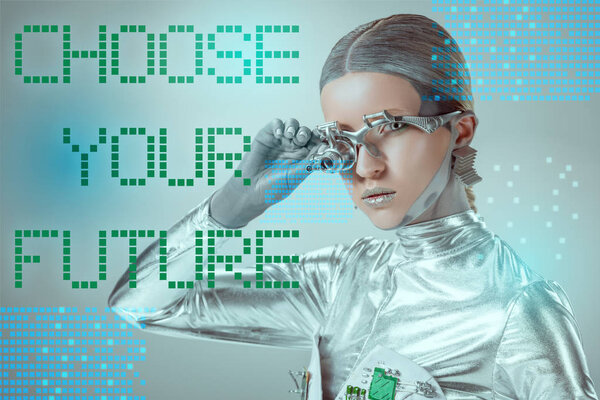 futuristic silver cyborg adjusting eye prosthesis and looking at camera on grey with "choose your future" lettering