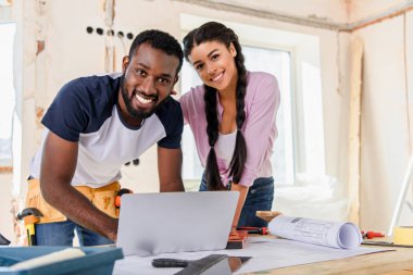smiling couple using laptop and looking at camera while making renovation of home clipart