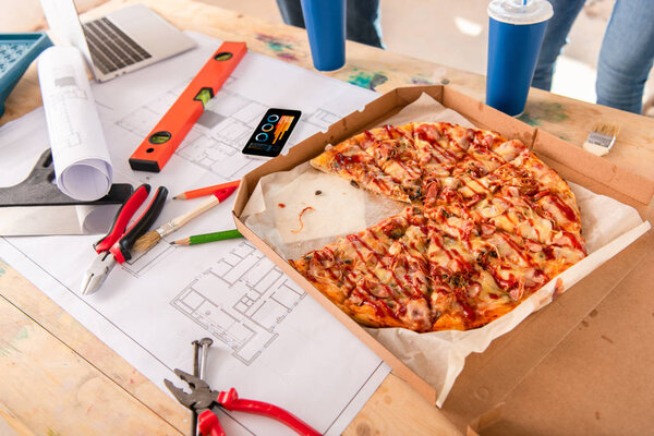 close-up shot of box with pizza, tools and smartphone with graphs on screen on building plan