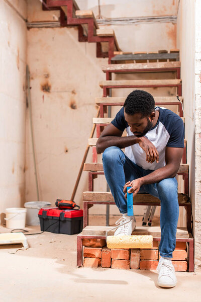 african american man sitting on stairs with roller brush during renovation of home