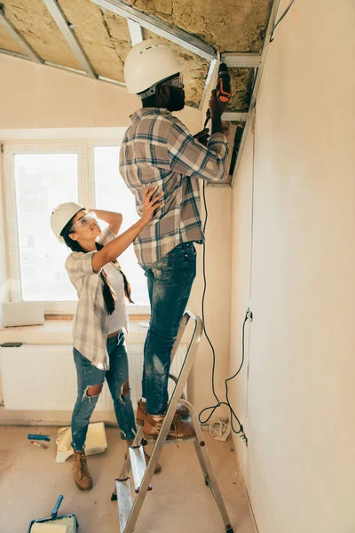 在装修房屋时 他的女朋友站在附近 戴着安全帽 戴着护目镜 在梯子上进行电钻的男子的侧视图 — 图库照片