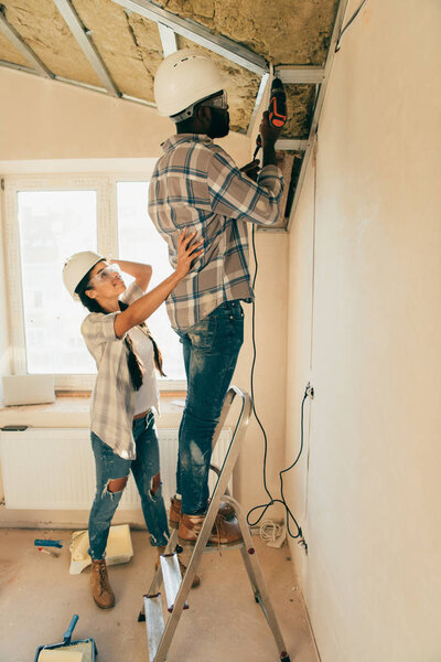 side view of man in hard hat and goggles working with power drill on ladder while his girlfriend standing near during renovation of home