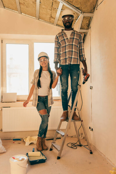 african american man in hard hat and goggles standing with power drill on ladder while his girlfriend standing near during renovation of home