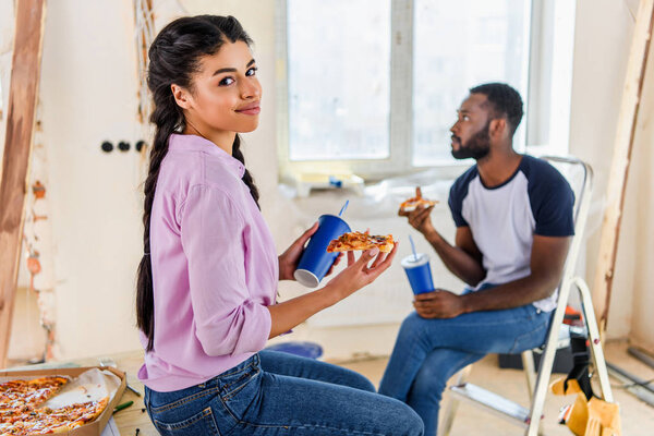 attractive african american woman with pizza and soda looking at camera while her boyfriend sitting behind at new home