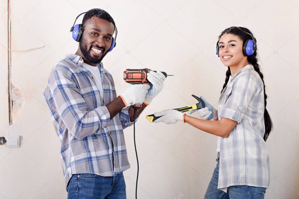 laughing young african american couple in protective headphones posing with drill and hammer during renovation of new home
