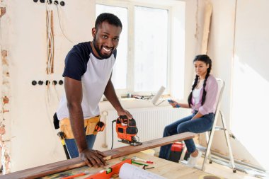 handsome african american man working with jigsaw while his girlfriend sitting near during renovation at home clipart