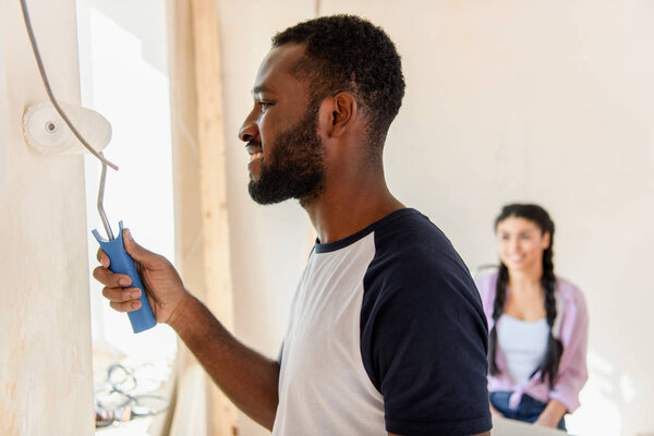 side view of happy african american man painting wall by paint roller while his girlfriend sitting behind at home