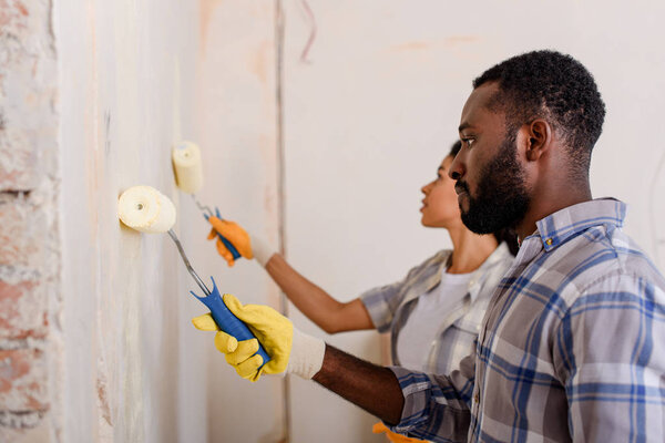 side view of african american couple painting wall by paint rollers at new home