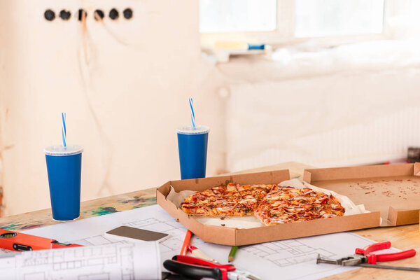 selective focus of pizza, soda, blueprint, tools and smartphone with blank screen on table