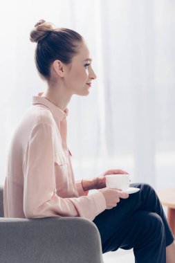 neşeli güzel iş kadını koltukta ofis, çay ile oturup yan görünüm