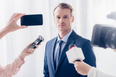 handsome businessman giving interview to journalists and looking at camera in office clipart