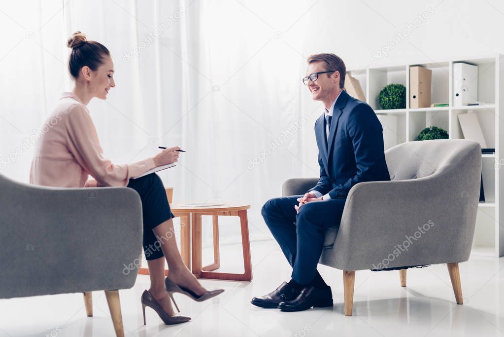side view of handsome smiling businessman giving interview to beautiful journalist at workspace