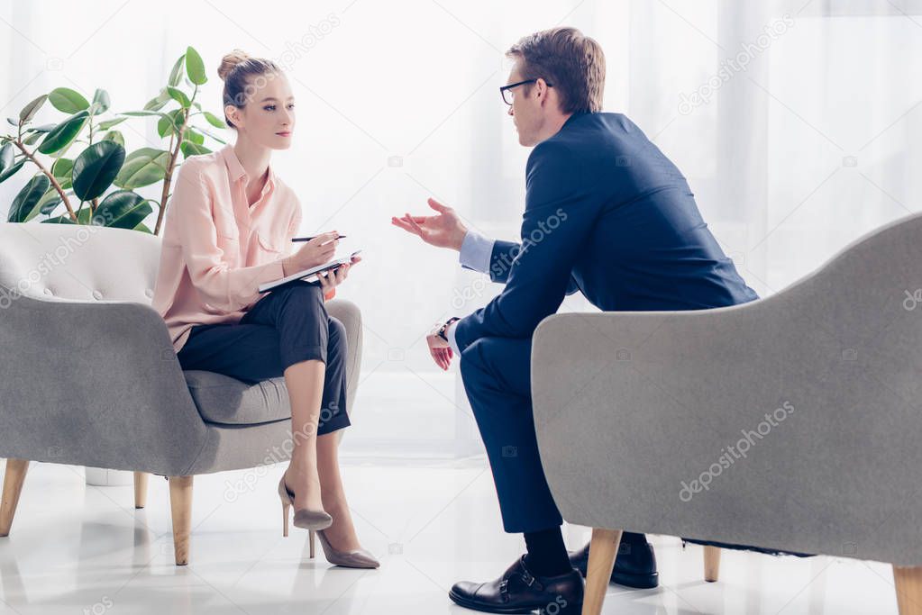 handsome businessman giving interview to journalist in office, they sitting in armchairs and looking at each other