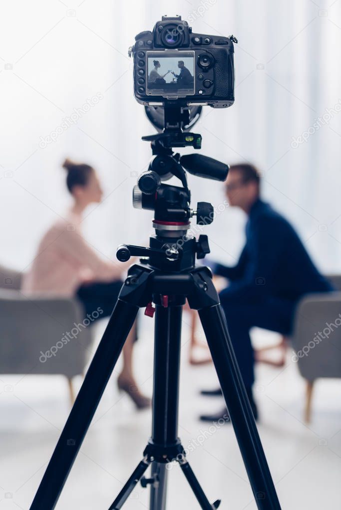side view of businessman giving interview to journalist in office, camera on tripod on foreground