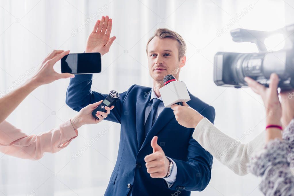 handsome businessman giving interview to journalists in office, waving hand and showing thumb up