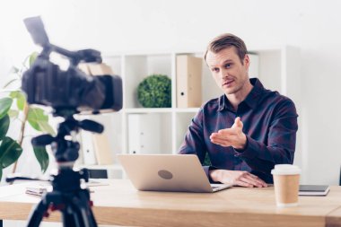 cheerful handsome male video blogger recording vlog and gesturing in office clipart