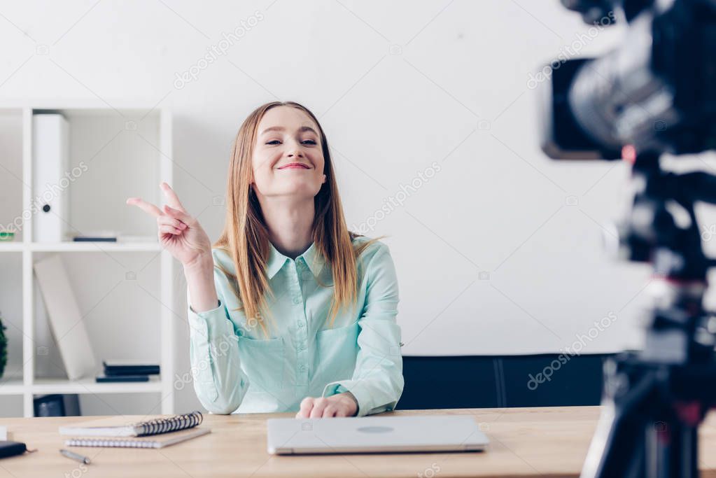 smiling attractive female video blogger recording vlog and showing two fingers in office