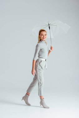 woman in grey clothes looking at camera and posing with transparent umbrella on grey background clipart