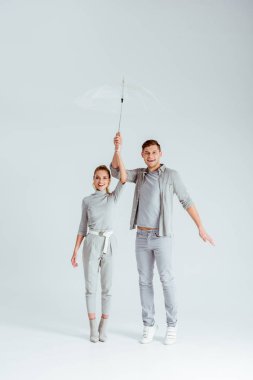 happy couple looking at camera, standing on tiptoe and posing with transparent umbrella on grey background clipart
