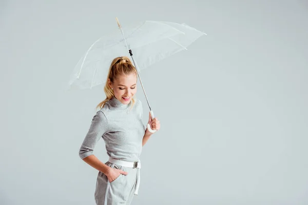 Улыбающаяся Женщина Серой Одежде Рука Кармане Позирует Прозрачным Зонтиком Изолированным — Бесплатное стоковое фото