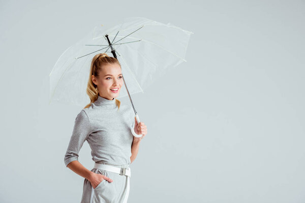 beautiful woman in grey clothes and hand in pocket posing with transparent umbrella isolated on grey