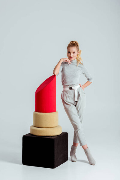 woman touching chin, posing near big red lipstick model and looking at camera on grey background