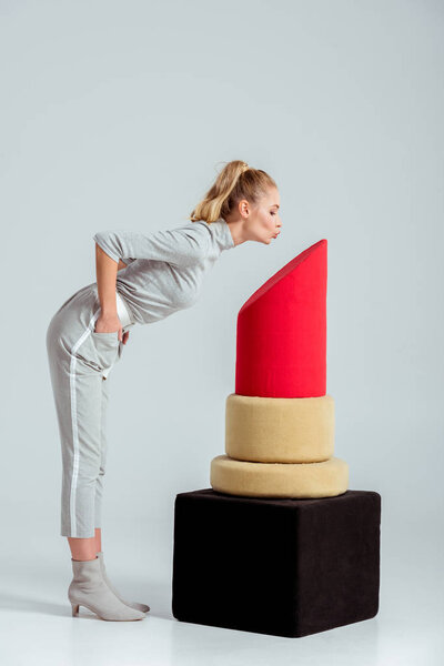 Woman giving air kiss near red lipstick model on grey background