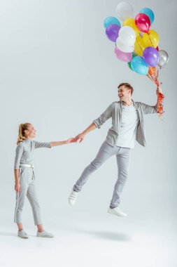 gri giyim gri arka plan üzerinde renkli balonlar bohça ile havada atlama mutlu adamın elini tutan kadın