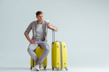 handsome man sitting on yellow suitcase and using smartphone on grey background, travel concept clipart