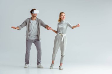 happy couple holding hands while man experiencing virtual reality on grey background clipart