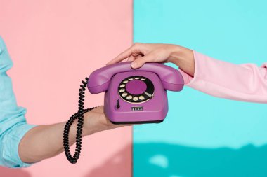 cropped view of man and woman holding purple vintage telephone on pink and blue background clipart