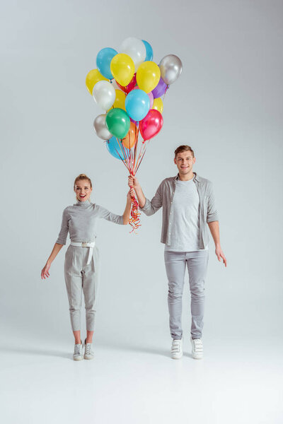 smiling couple standing on tiptoe, looking at camera and holding bundle of colorful balloons on grey background
