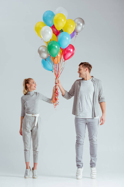 couple standing on tiptoe, looking at each other and holding bundle of colorful balloons on grey background