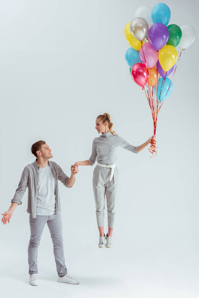 handsome man holding hand of woman jumping in air with bundle of colorful balloons on grey background