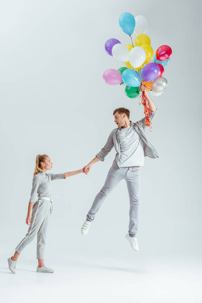 beautiful woman holding hand of man jumping in air with bundle of colorful balloons on grey background