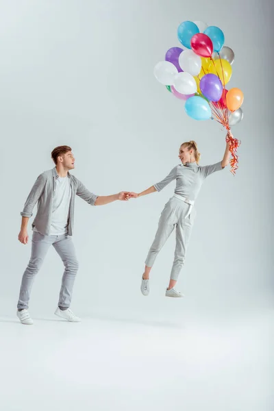 man holding hand of woman jumping in air with bundle of colorful balloons on grey background