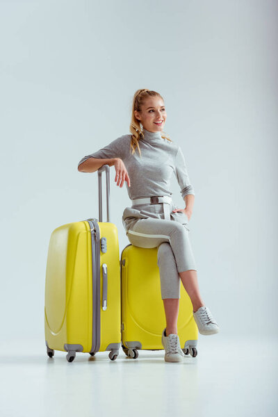 smiling woman sitting on suitcases with crossed legs on grey background, travel concept