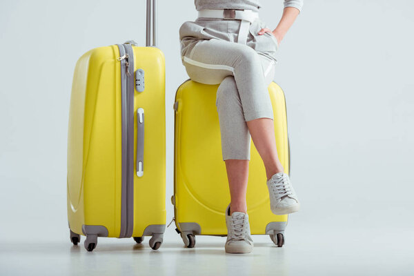 cropped view of woman sitting on suitcases with crossed legs on grey background, travel concept