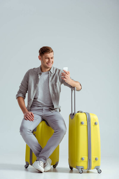 smiling man sitting on yellow suitcase and using smartphone on grey background, travel concept