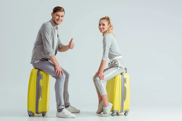 夫妇坐在行李箱上 而男子显示拇指在灰色背景上签名 旅行概念 — 图库照片