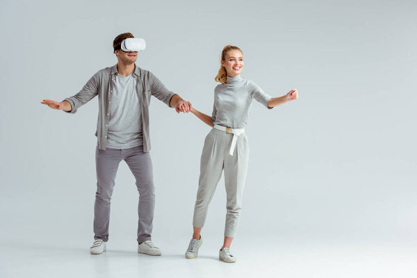 happy couple holding hands while man experiencing virtual reality on grey background