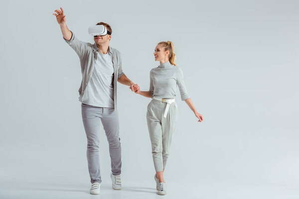 couple holding hands while man experiencing virtual reality and gesturing on grey background