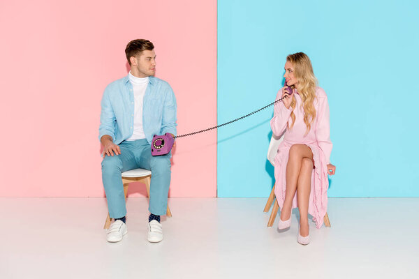 smiling woman having conversation while man holding purple vintage telephone on pink and blue background