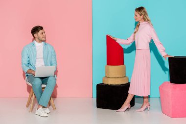 oje ve ruj modelleriyle sandalyede oturan ve pembe ve mavi arka plan üzerinde dizüstü kullanarak adam kılığında kadın