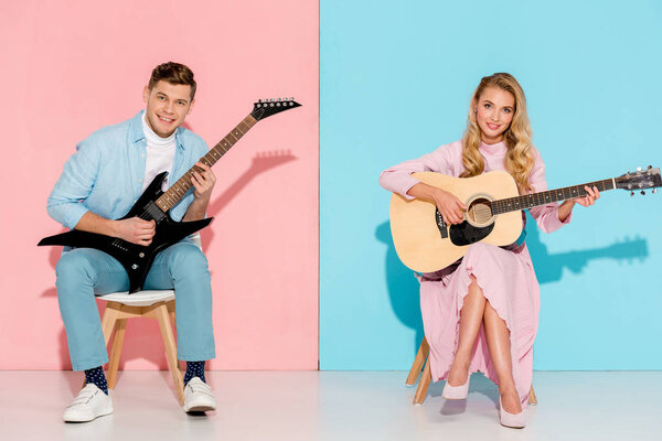 couple sitting on chairs, playing electric and acoustic guitars and looking at camera on pink and blue background