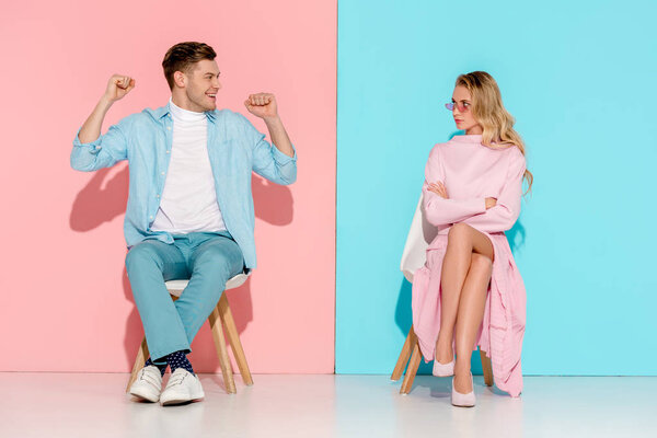 dissatisfied woman with arms crossed and cheering man with clenched fists sitting on chairs on pink and blue background