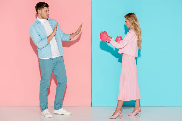 scared man gesturing with hands near aggressive woman in pink boxing gloves on pink and blue background