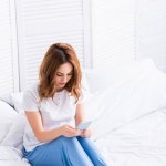 Attraktiv kvinna med ingefära hår vila i vit säng och med smartphone hemma på helgen