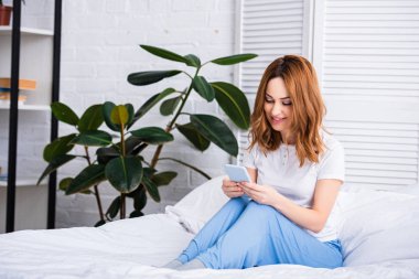 attractive woman with ginger hair resting in bed and using smartphone at home on weekend clipart